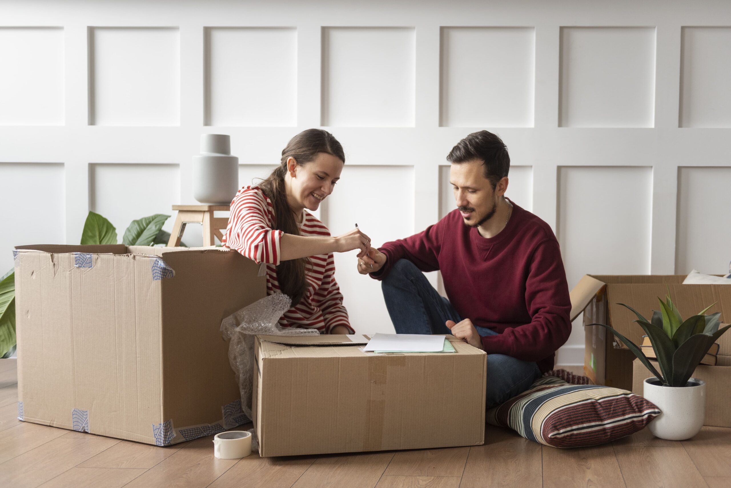 Deux personnes souriantes scellent un accord avec une poignée de main au milieu de cartons, meubles et plantes, pendant le déménagement. check-list déménagement entreprise plausible.