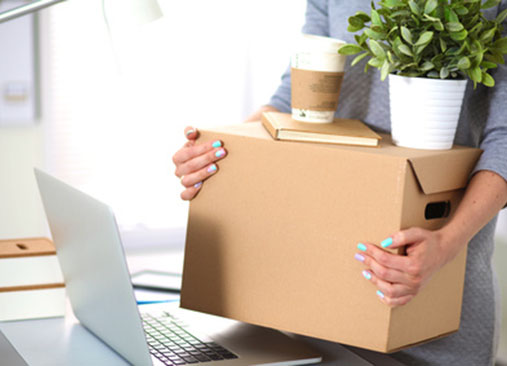 Personne tenant une boîte en carton près d’un ordinateur portable, avec une plante et des livres sur le bureau.