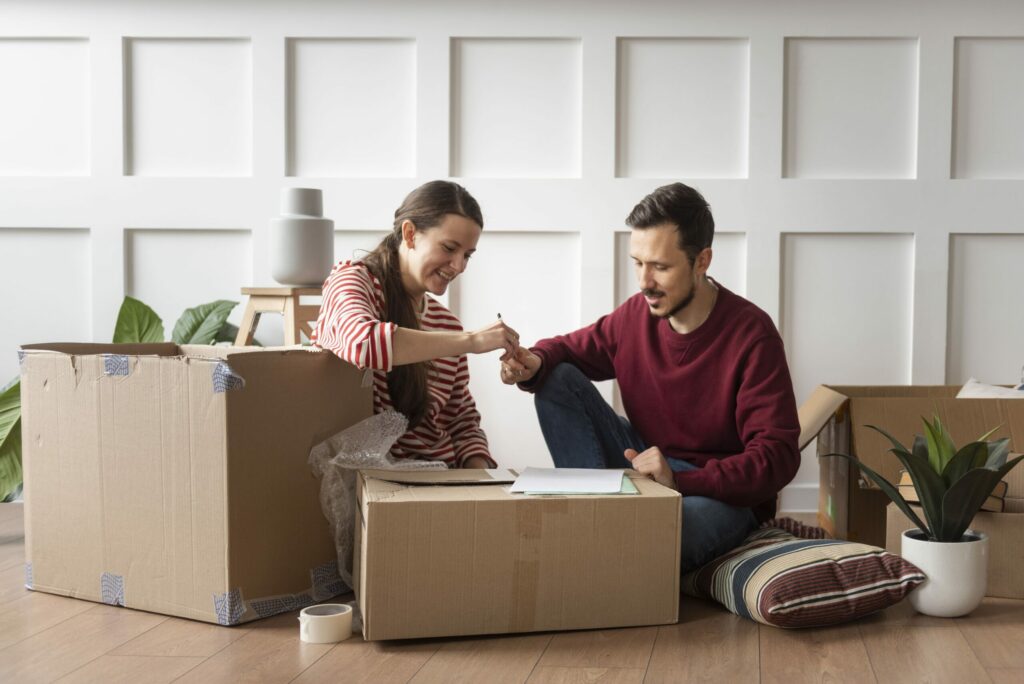 Deux personnes fêtent autour d’un déménagement : ils se serrent la main au-dessus d’une grande boîte en carton, dans un salon, entourés de cartons et de plantes.